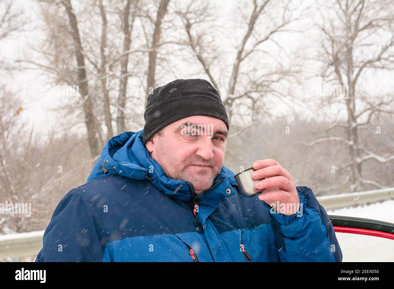 Mann mittleren Alters trinkt Tee aus Eisen Thermoskanne im Winter. Schneefall und Äste im Raureif. Ruhen Sie sich auf dem Weg in der Kälte aus. Stockfoto