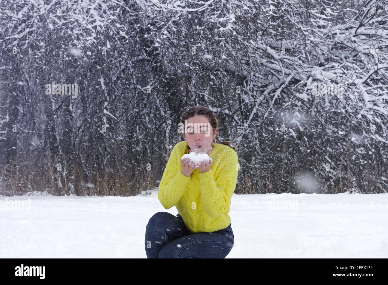 Schöne junge Frau bläst Schnee aus den Händen im Winterwald. Pulverschneemflocken fliegen hoch. Fröhliche Dame in leuchtend gelben Jacke vor Hintergrund von Whi Stockfoto