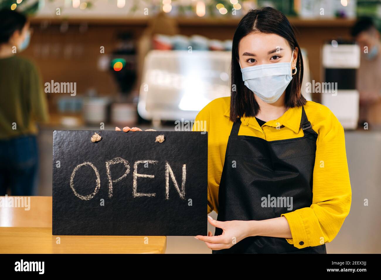 Herzlich Willkommen. Die junge asiatische Kellnerin steht im Innenbereich eines Restaurants oder Cafés mit einer schützenden medizinischen Maske und einer schwarzen Schürze und hält das Schild OFFEN. Unterstützung des Konzepts für kleine Unternehmen Stockfoto