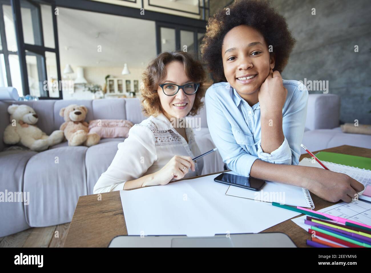 Fröhliche gemischte Rasse teen Mädchen und weibliche Lehrer lächelnd an Kamera während Hausaufgaben zusammen im Wohnzimmer zu tun Zu Hause Stockfoto