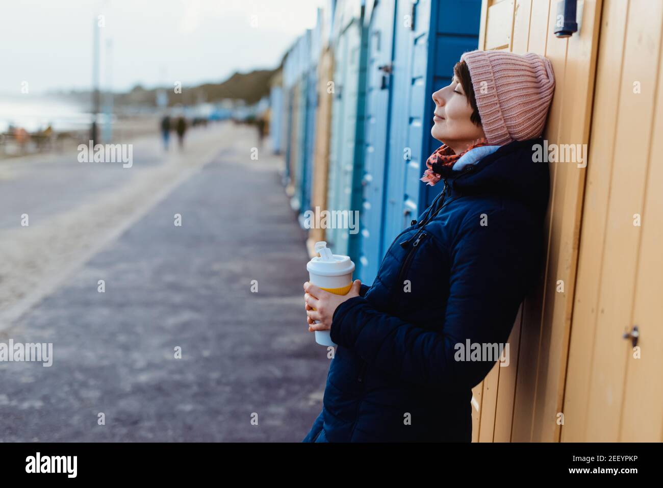 Lächelnde Frau mit geschlossenen Augen in warmer Kleidung mit einer wiederverwendbaren Tasse mit einem heißen Getränk in der Nähe von bunten Strandbuden und genießen den Moment. Einfach Stockfoto