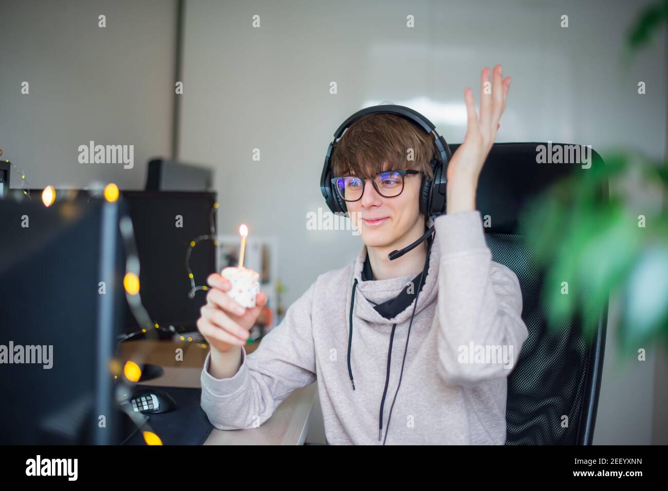 Junger Mann mit festlichem Cupcake mit brennender Kerze auf virtueller Geburtstagsparty während des Lockdown. Generation Z Mann feiert seinen Geburtstag allein auf Stockfoto