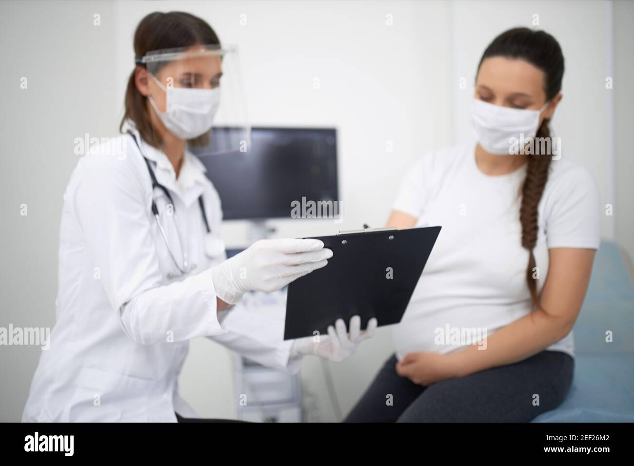 Junge Frau in Erwartung in der medizinischen Maske mit regelmäßigen Untersuchungen im modernen Krankenhaus. Gynäkologe trägt eine schützende Gesichtsmaske bei der Bereitstellung von d Stockfoto