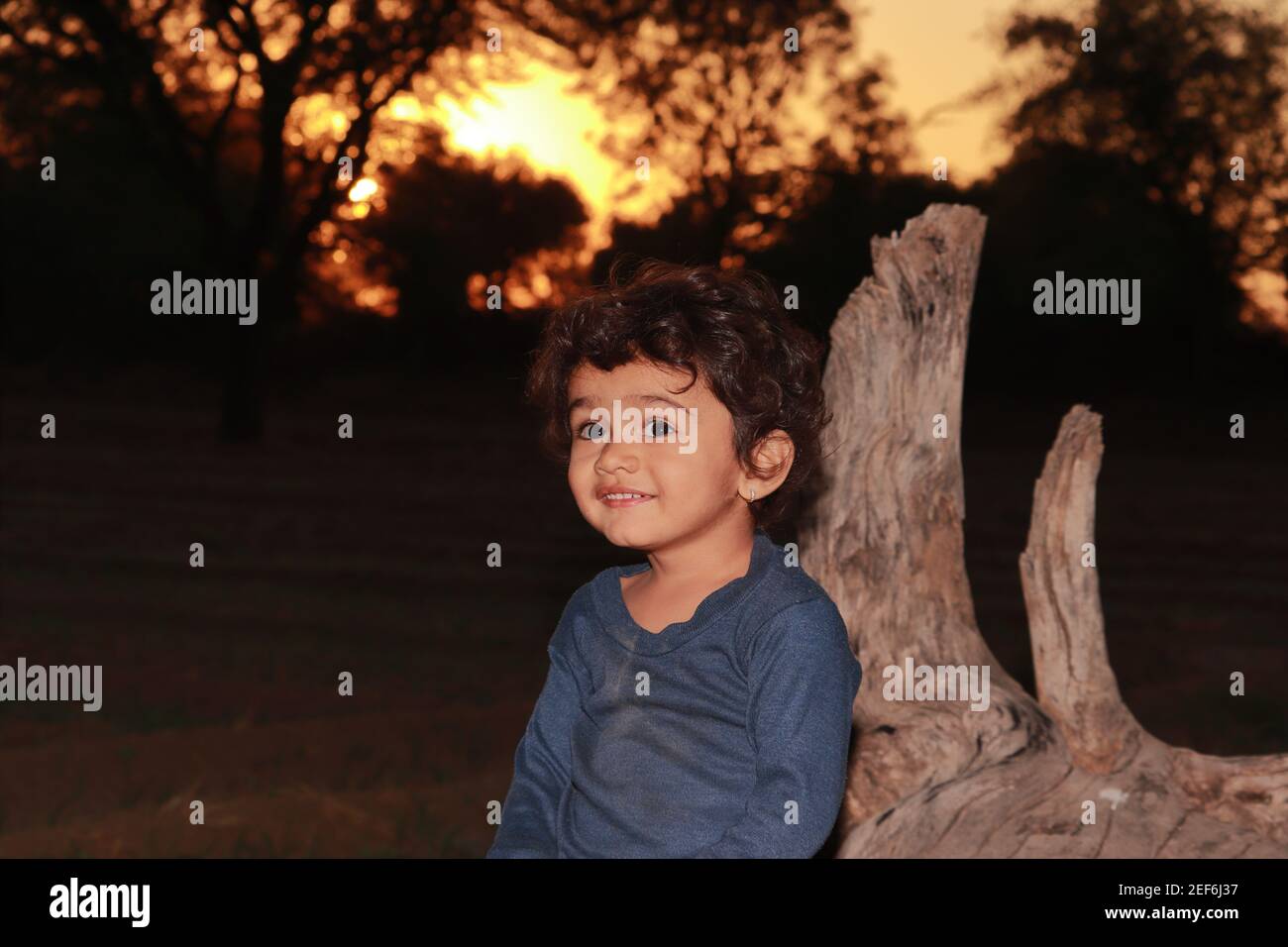 Ein schöner kleiner indischer Junge Kind, sitzt auf einem trockenen Holz im Garten bei Sonnenuntergang, indischer Junge Kind lächelt Blick auf die Kamera. Konzept für Sweet c Stockfoto
