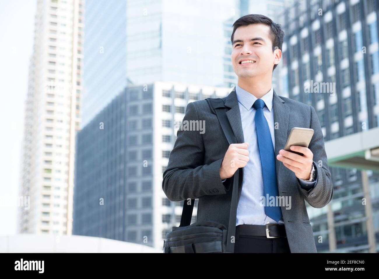 Lächelnder Geschäftsmann, der sein Mobiltelefon benutzt, während er seine Tasche trägt und zu Fuß geht In der Stadt - Geschäftsreisen und mobile Roaming-Konzepte Stockfoto