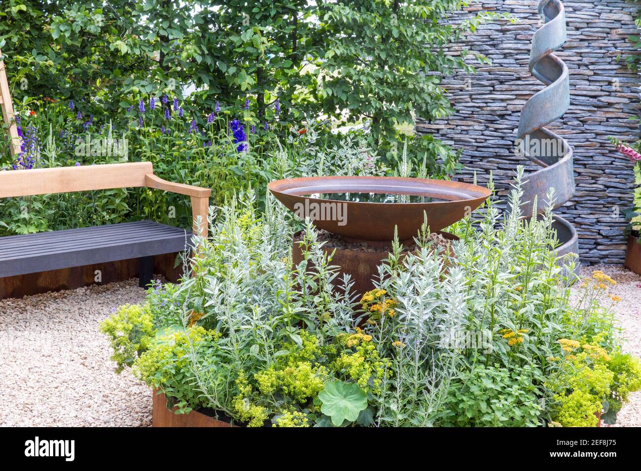 Kleiner englischer Hofgarten mit Spiralskulptur - Kortenstahl Wasserspiel - Hecken - Schotterweg mit Gartenbank Flower Borders England Großbritannien Stockfoto