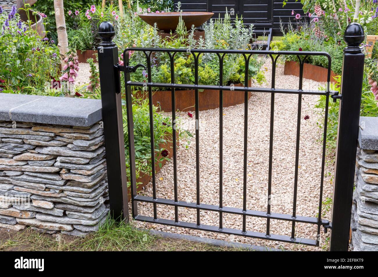 Ein bemaltes schwarzes Metallgartentor - trockene Steinmauer Blick auf englischen Cottage Country Garten mit Schotterweg und Blumenbeete Garten Grenze England Großbritannien Stockfoto