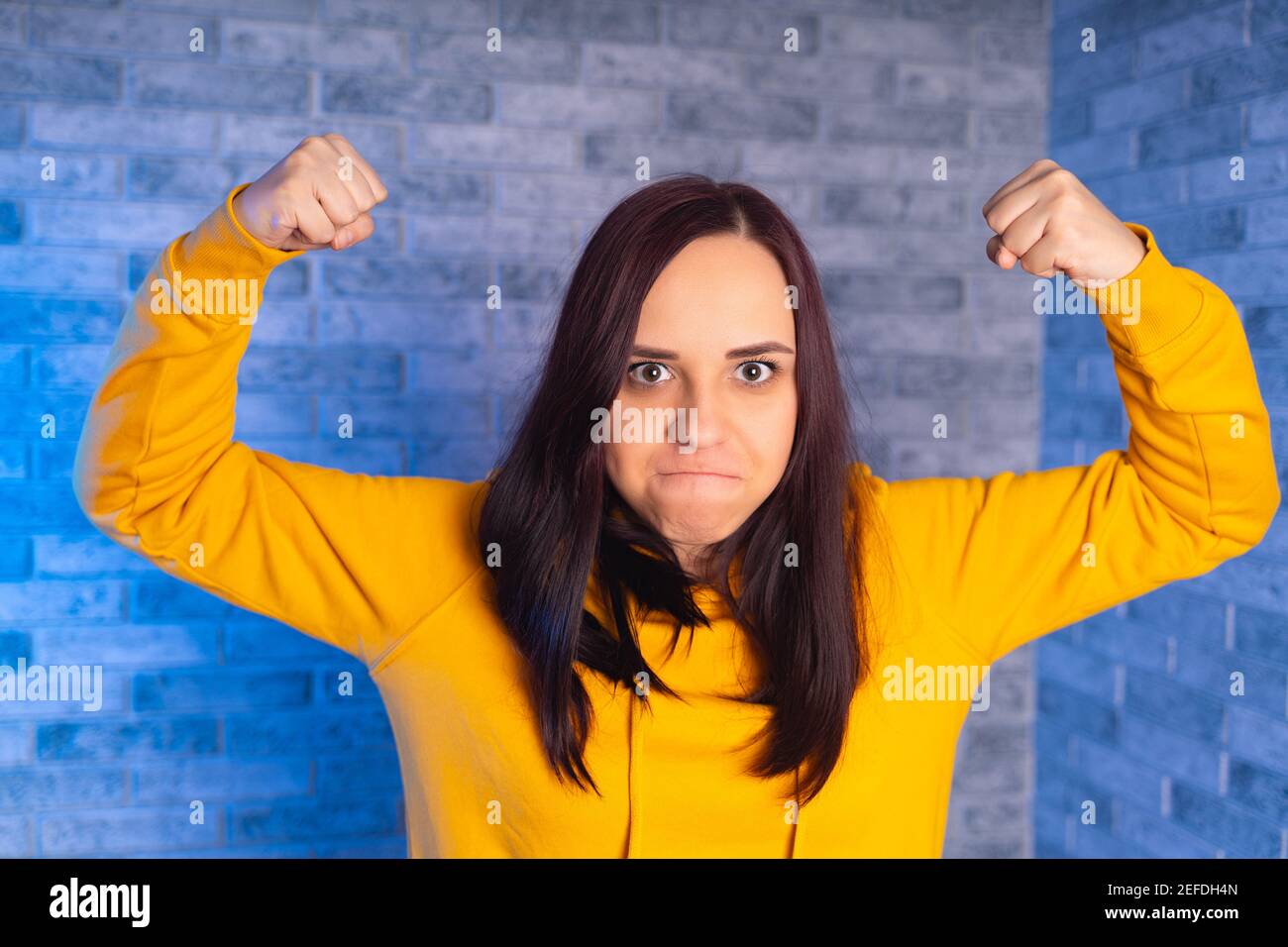Porträt einer jungen Frau, die mit ihren Händen Kraft zeigt. Nahaufnahme der unzufriedenen Frau in gelben Hoodie zeigt Bizeps auf grauem Backstein Hintergrund Stockfoto