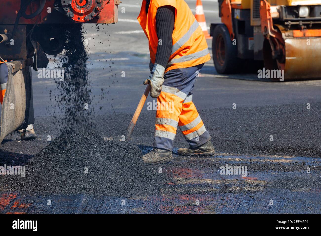 Ein Straßenarbeiter in orangefarbenen Overalls renoviert einen Abschnitt der Straße mit heißem Asphalt vor dem Hintergrund einer Straßenwalze in Unschärfe. Konzept der Straßenreparatur Stockfoto