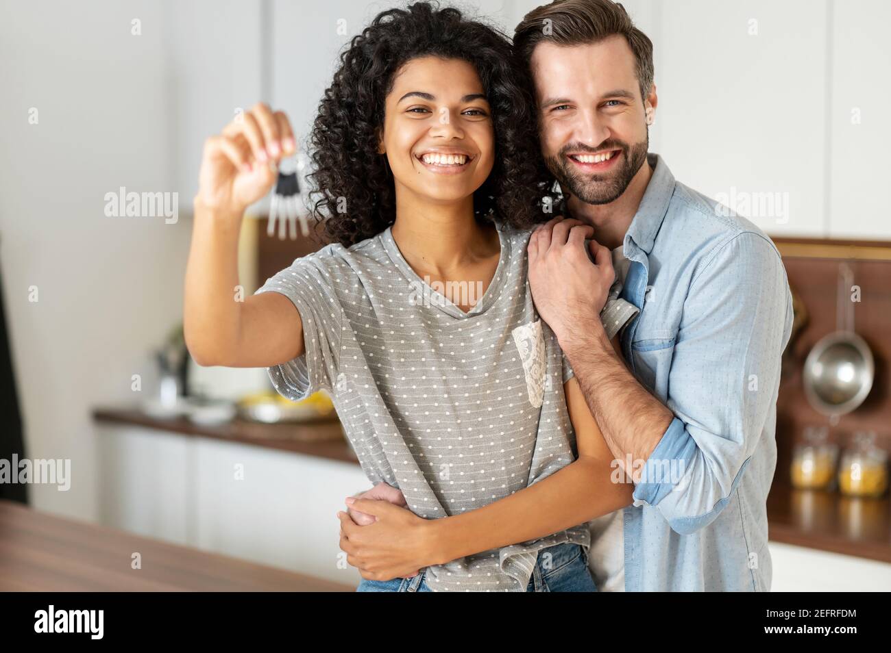 Fröhliche junge multi ethnischen Paar in der Liebe stehen in umarmt die Küche, hält die Schlüssel eines neuen Hauses. Freudige internationale Neuvermählte sind neu eingezogen Stockfoto