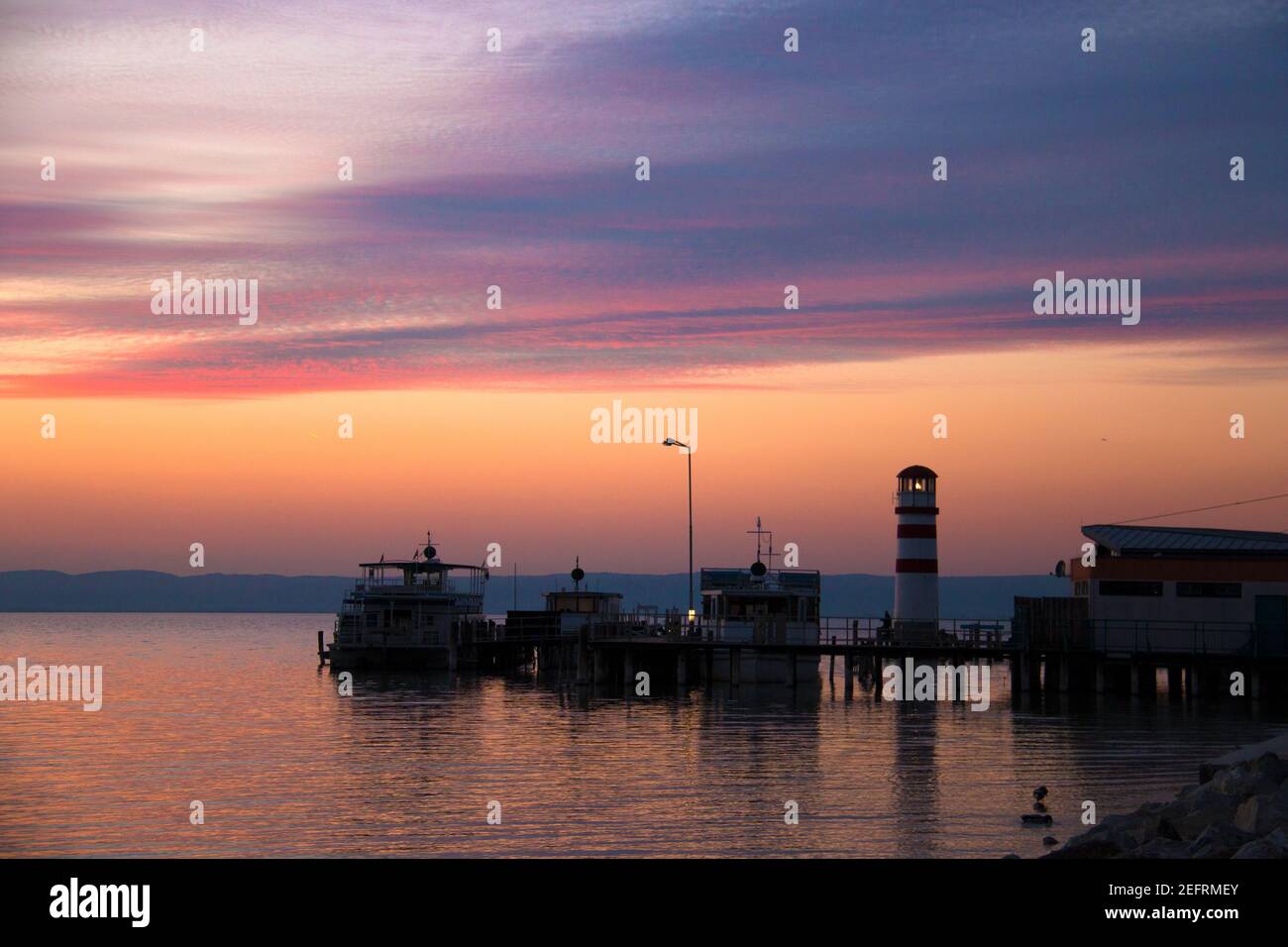 Schöner Dämmerungshimmel nach Sonnenuntergang über dem Meerwasser mit Blick auf angedockte Boote auf Pier und Leuchtturm Turm mit Schatten Reflexion auf dem Wasser, ora Stockfoto