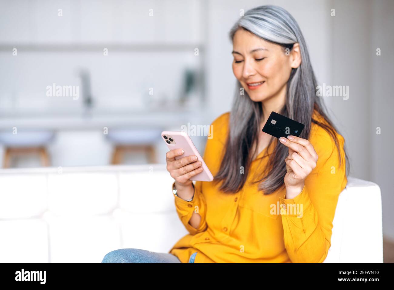 Nahaufnahme eine Kreditkarte und ein Handy in weiblichen Händen. Defokussierte asiatische grauhaarige Frau mittleren Alters verwendet eine Kreditkarte und Smartphone, kauft und zahlt online, Internet-Banking-Konzept Stockfoto