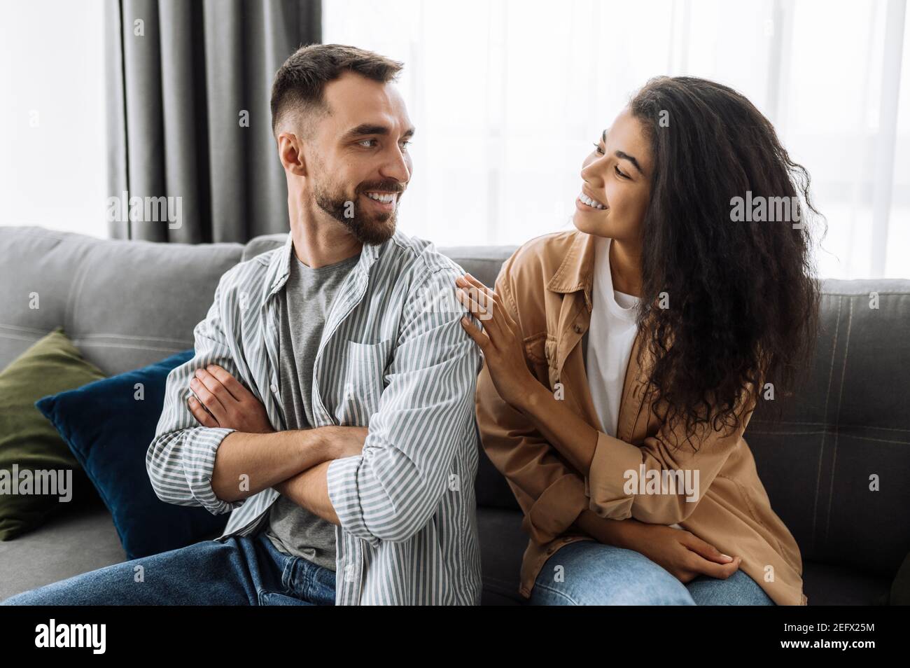 Fröhliches Ehepaar, das zu Hause chillt und auf dem Sofa sitzt. Kaukasischen Mann schaut mit Liebe auf ihre schöne afroamerikanische Frau, Wochenende zusammen Stockfoto