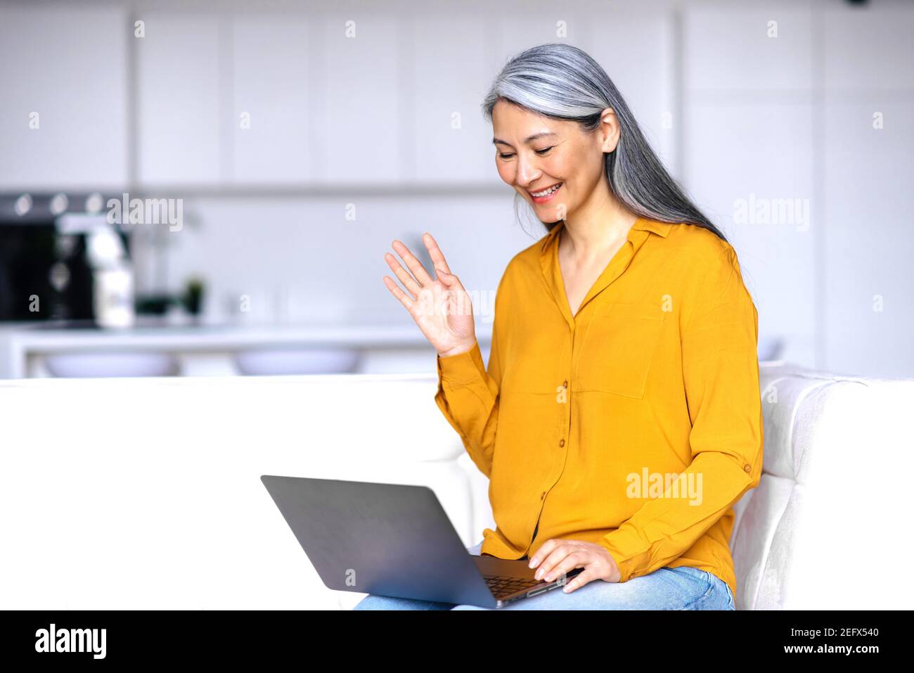 Lächelnd freundliche asiatische grauhaarige ältere Dame, sitzt auf der Couch im Wohnzimmer, mit Laptop, während der Arbeit aus der Ferne, kommuniziert mit Geschäftskollegen oder Freunden, Wellen Hand, Grüße Stockfoto