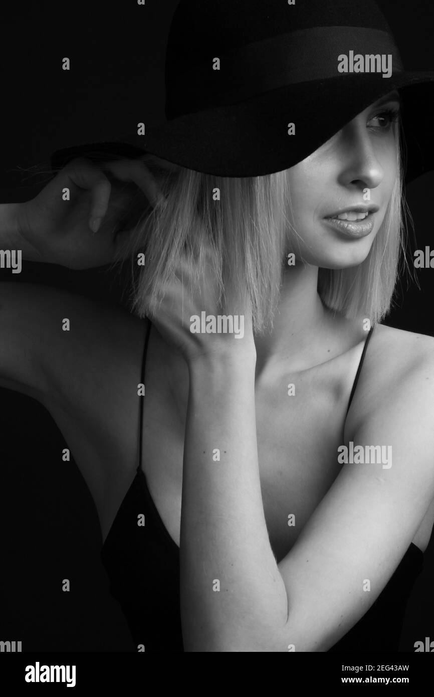 Schwarz-weiße Portrait Blondinen auf schwarzem Hintergrund in einem Hut und Tank Top mit dünnen Trägern. Hände am Ohr, passt den Ohrring Stockfoto
