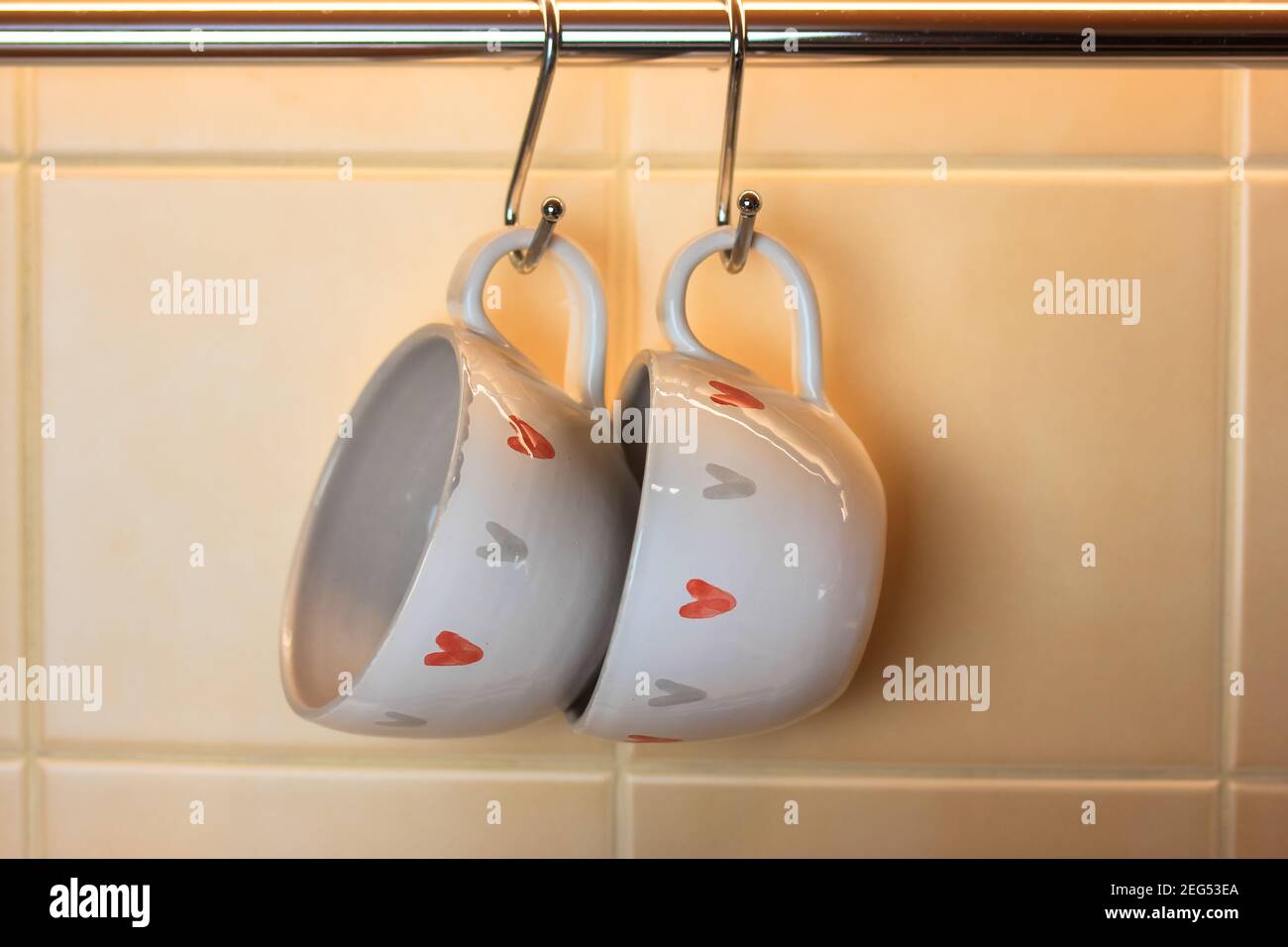 Zwei Kaffeetassen mit dem Bild von Herzen hängen an Haken über einem Tisch in der Küche. Leerzeichen für Text. Valentinstag-Konzept Stockfoto