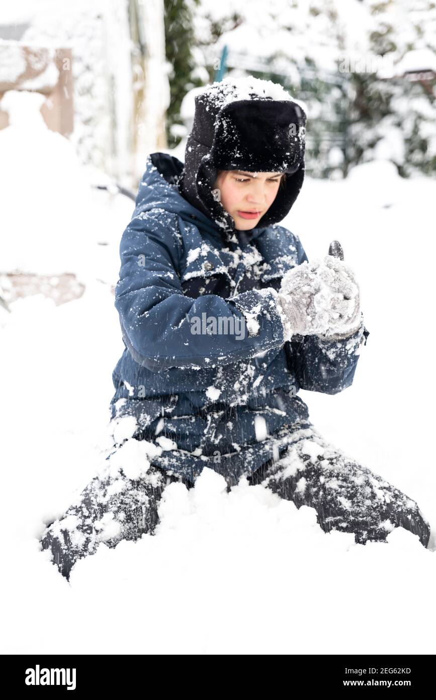 Niedlichen Jungen in Ohrenklappen Hut sitzen im Schnee und machen Schneebälle. Teenager haben Spaß und genießen Winterferien. Selektive Fokussierung und Tonung Stockfoto