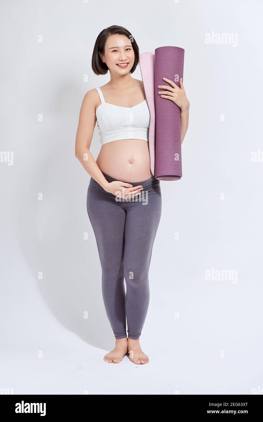 Junge attraktive schwangere Frau berühren ihren Bauch, während Yoga-Matte in den Händen halten, schwangere Mutter tun körperliche Übungen, posiert isoliert über w Stockfoto