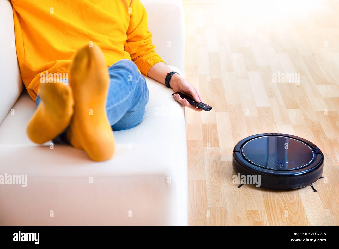 Mann ruht sich aus, während der Roboter-Staubsauger Aufgaben erledigt, zu Hause sauber arbeitet. Man steuert einen Roboter-Staubsauger mit einer Fernbedienung. Reinigungsroboter V Stockfoto