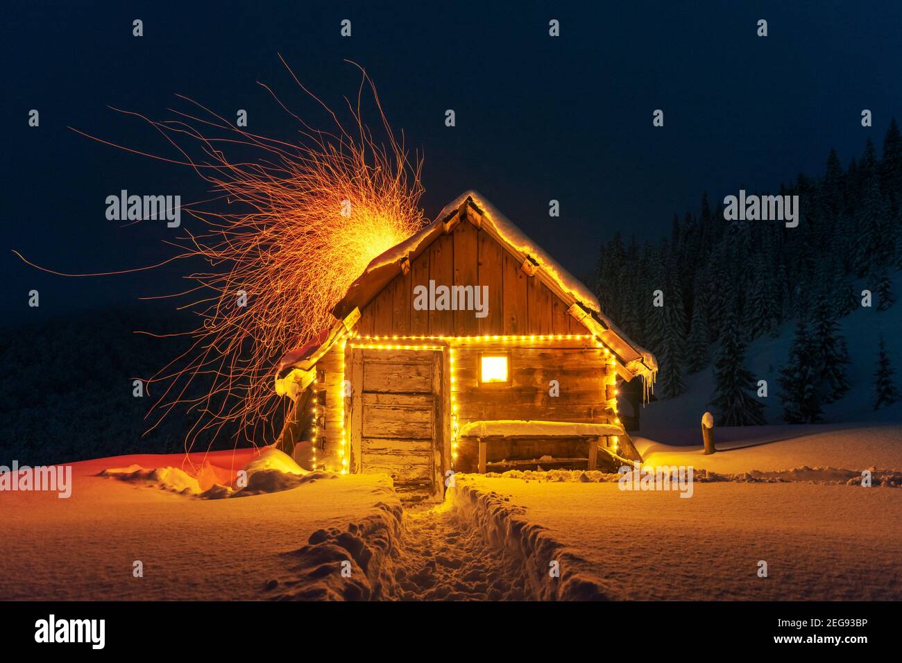 Fantastische Winterlandschaft mit glühender Holzhütte im verschneiten Wald. Feuerfunken fliegen aus dem Kamin. Weihnachtsfeiertagskonzept Stockfoto