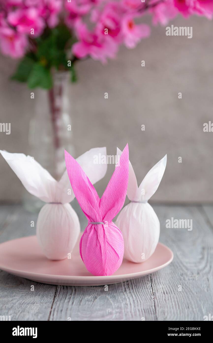Drei Eier Hase in der farbigen Papierverpackung auf dem Teller, daneben Zweig der Kirschblüten. Der Osterurlaub steht auf dem Tisch Stockfoto