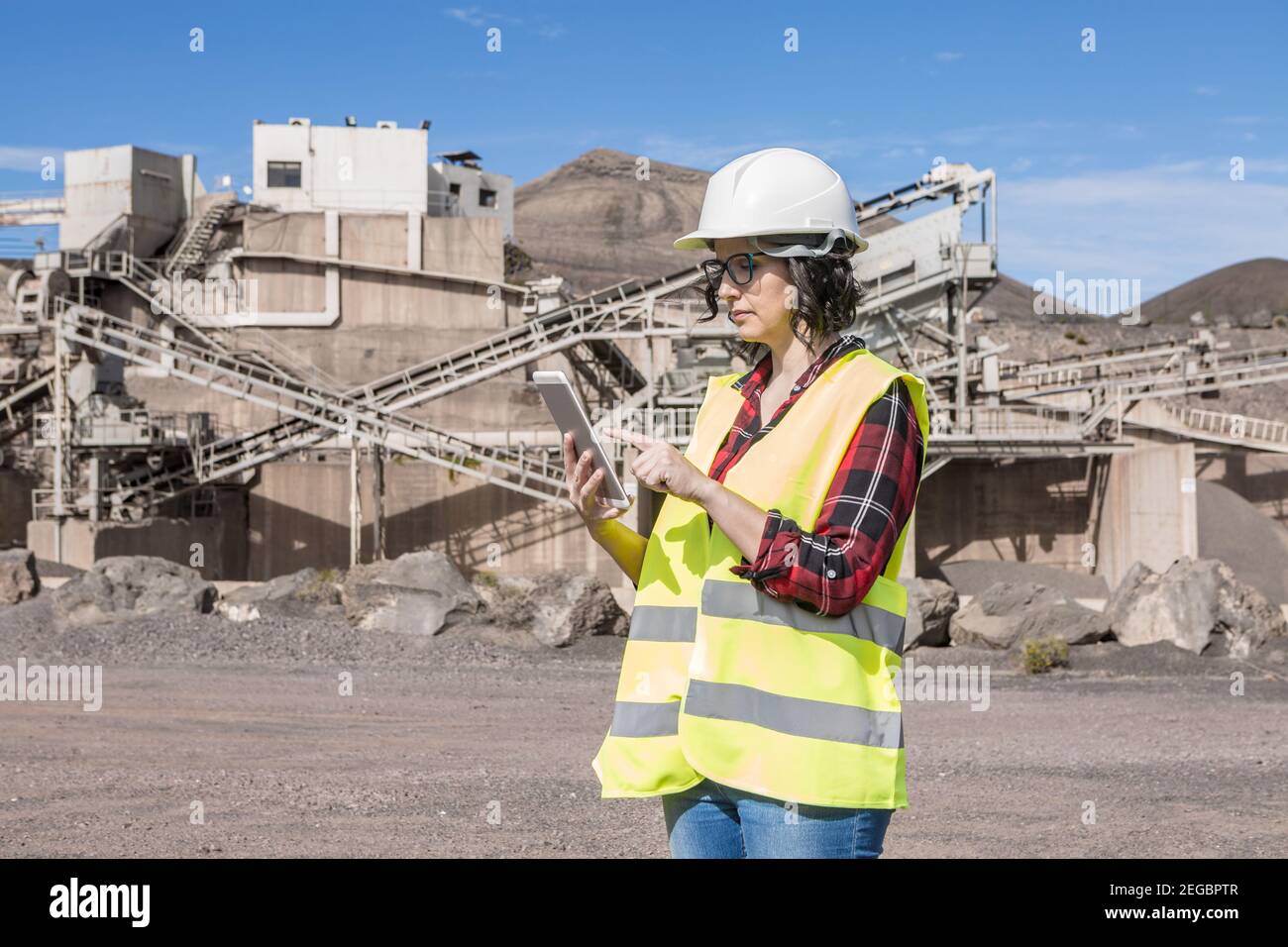 Seitenansicht der fokussierten Ingenieurin in Hardhut und Weste Prüfen von Informationen auf dem Tablet, während Sie in der Nähe einer Industrieanlage von stehen Konstruktion s Stockfoto