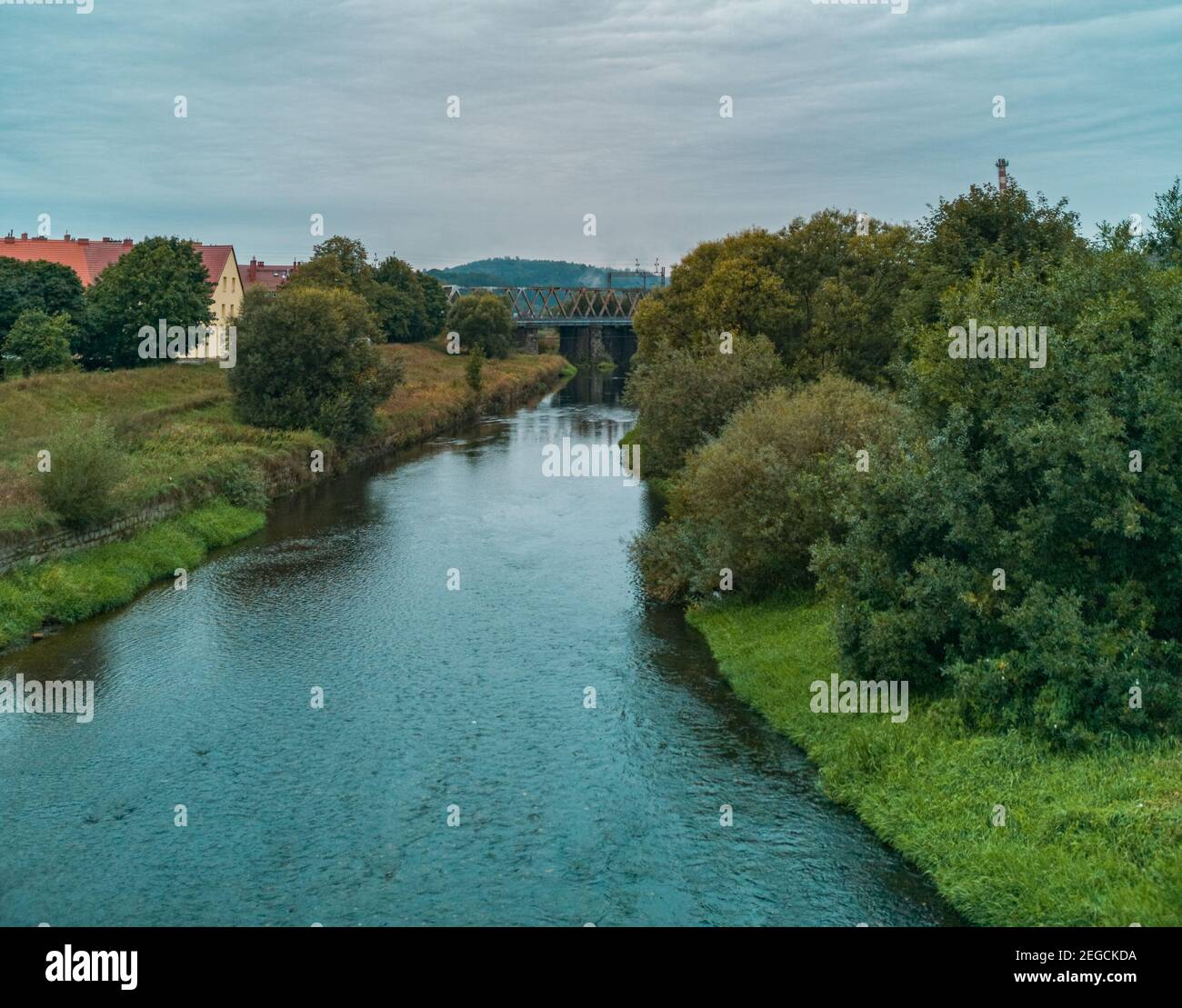 Wolkige Landschaft des kleinen Flusses zwischen Büschen und Bäumen mit Zugbrücke darüber Stockfoto