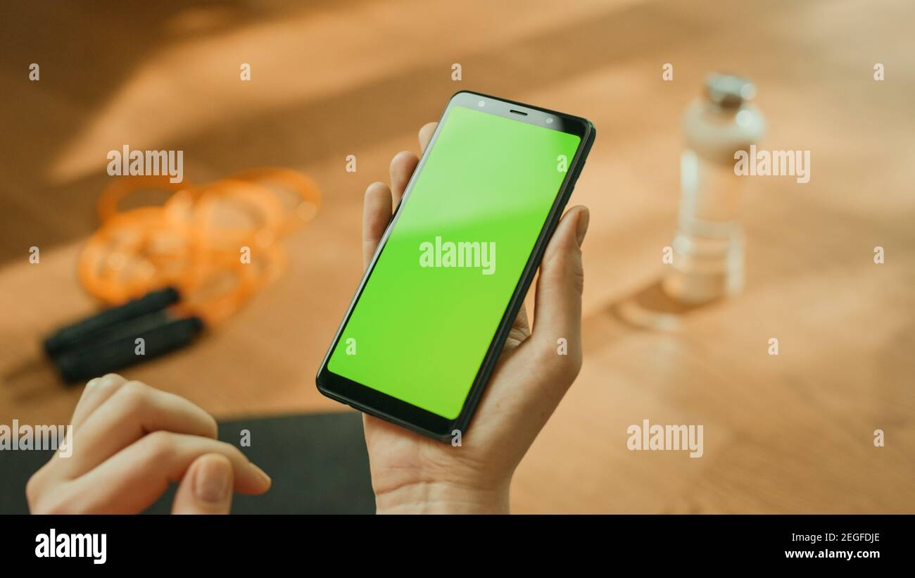 Fitness-Konzept: Frau Hält Chroma Key Green Screen Smartphone. Im Hintergrund Fitness-Matte, Springseil und Ausrüstung. Nahaufnahme des Betrachtungspunkts Stockfoto