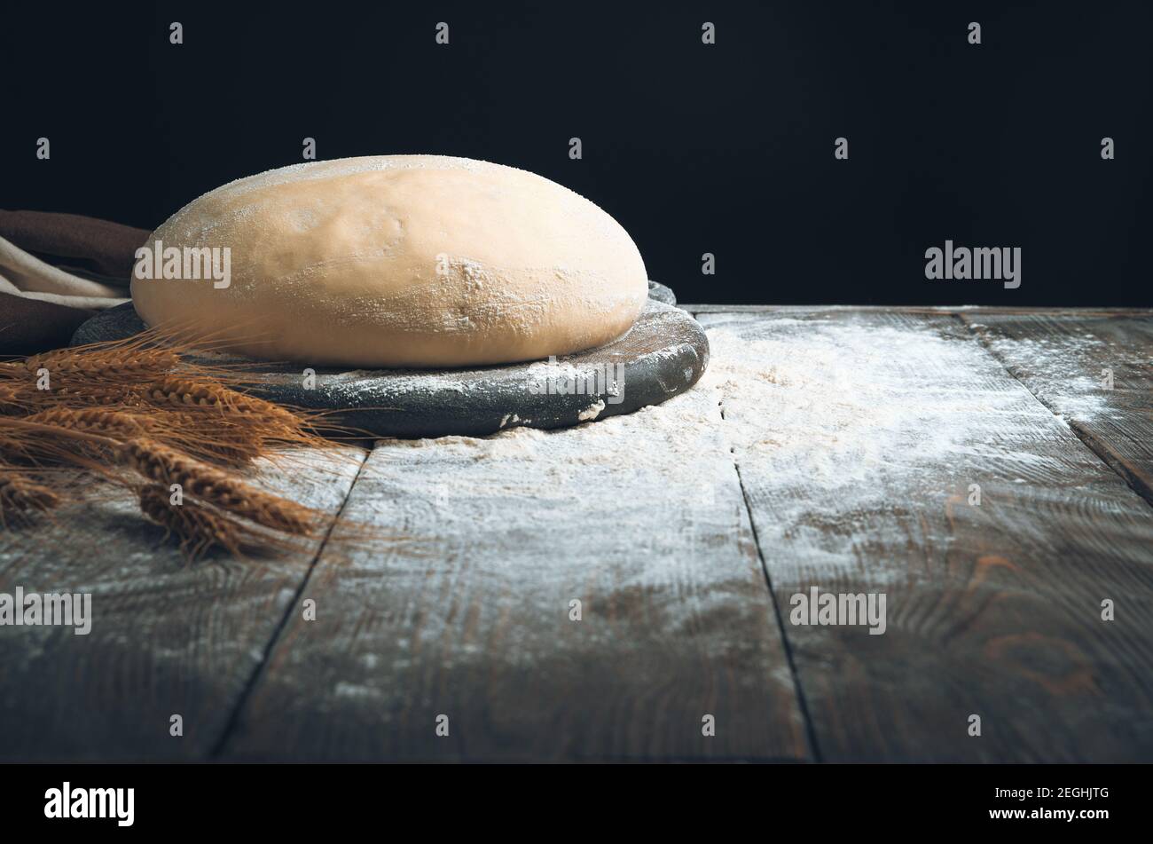 Runder Teig auf einem Brett in Mehl und Weizen auf dunklem Grund. Stockfoto