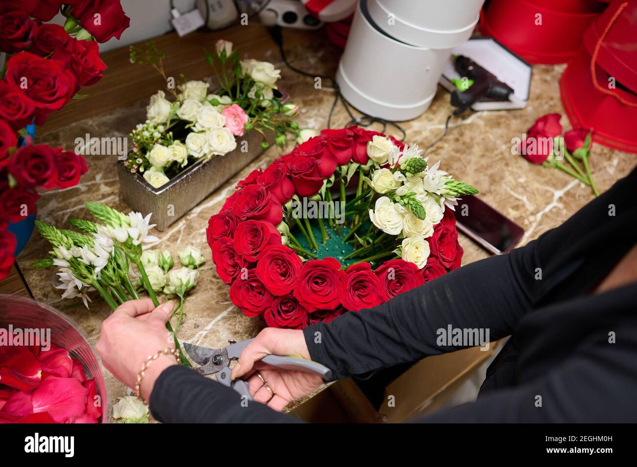 Blumenhändler, der Blumenbox mit roten Rosen und weißen Blumen auf dem Tisch macht, rote Rosen im Tank auf dem Hintergrund Stockfoto