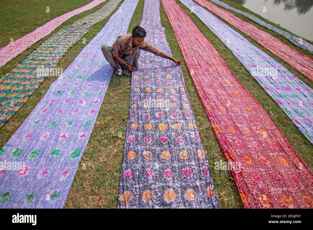 Narayanganj, Bangladesch. Februar 2021, 18th. Arbeiter trocknen Batik-Kleidung unter der Sonne auf einem offenen Feld als Teil des Prozesses der Schaffung traditioneller Batik. In dieser Technik wird Wachs verwendet, um atemberaubende Muster in großem Maßstab zu erstellen. Das Wachs verhindert, dass der Farbstoff in den Bereich eindringt, auf dem er platziert wird, so dass die Arbeiter erstaunlich komplexe mehrfarbige Designs erstellen können. Dann weichen sie die Tücher in einer sterbenden Emulsion ein, bevor sie die großen Baumwoll-, Seide- oder Wolltücher in der heißen Sonne zum Trocknen aufrollen. (Foto von Joy Saha/Pacific Press) Quelle: Pacific Press Media Production Corp./Alamy Live News Stockfoto