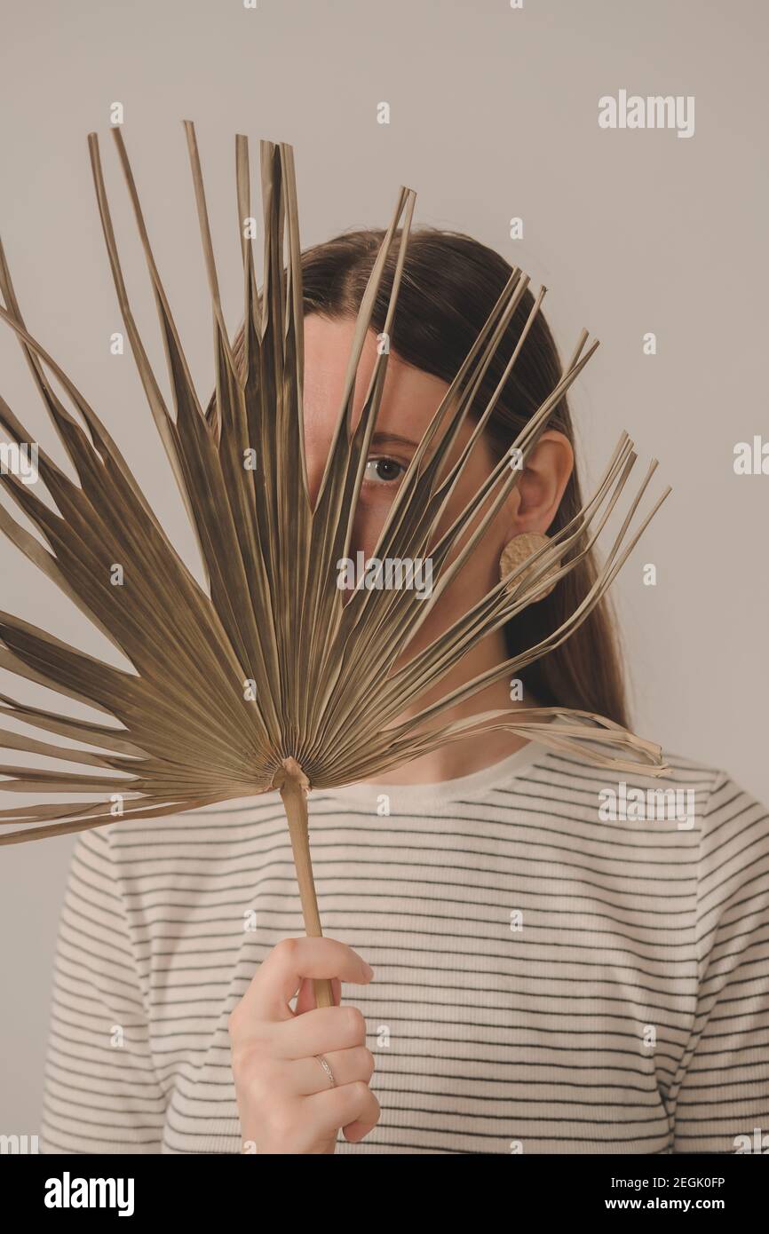Porträt einer Frau, die durch ein Palmblatt schaut. Junge kaukasische weibliche Person posiert mit getrockneten Blatt, neutrale und verblasste Farben Stockfoto