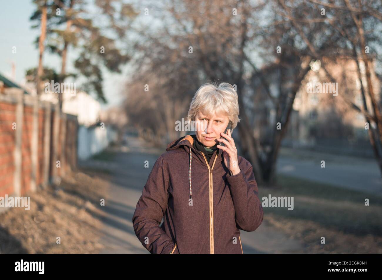 Eine schöne Frau mittleren Alters in legerer Kleidung spricht am Telefon mit einem Lächeln auf ihrem Gesicht. Abends in der Sonne durch die Stadt spazieren. Stockfoto