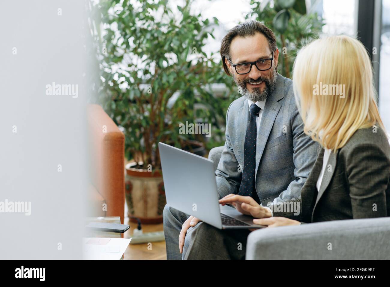 Erfolgreiche Kollegen mittleren Alters sitzen auf der Couch im Büro und diskutieren neue Businessplan. Schöner Mann mit blonden Haaren Frau mit Computer, über Ideen sprechen, Partnerschaft Konzept Stockfoto