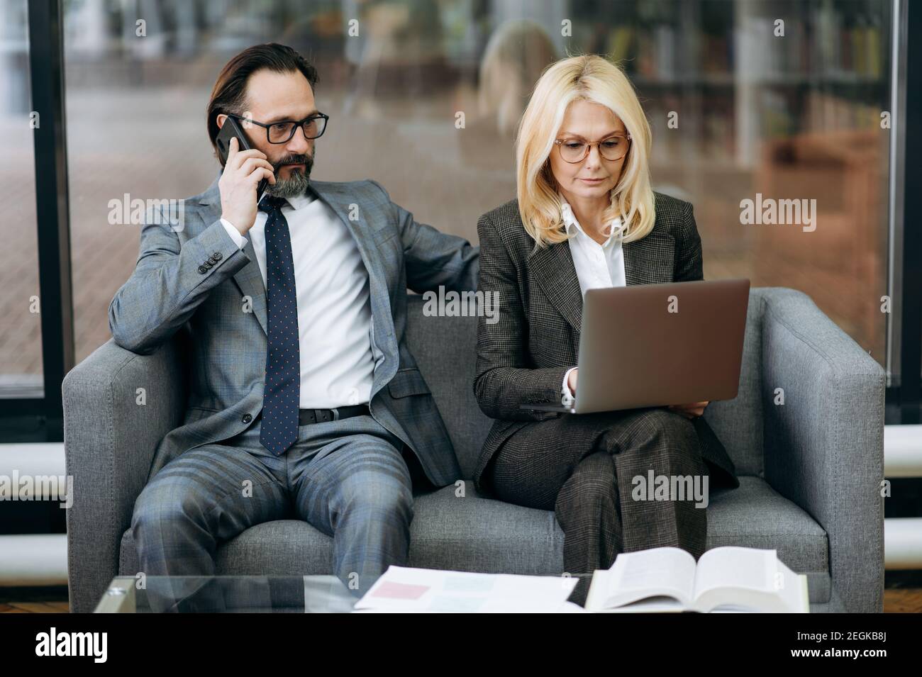 Seriöse Geschäftspartner sitzen auf dem Sofa im modernen Büro, mit Laptop und Smartphone. Reife Kollegen arbeiten an Unternehmensprojekten, Diskussionsideen, Partnerschaftskonzept Stockfoto