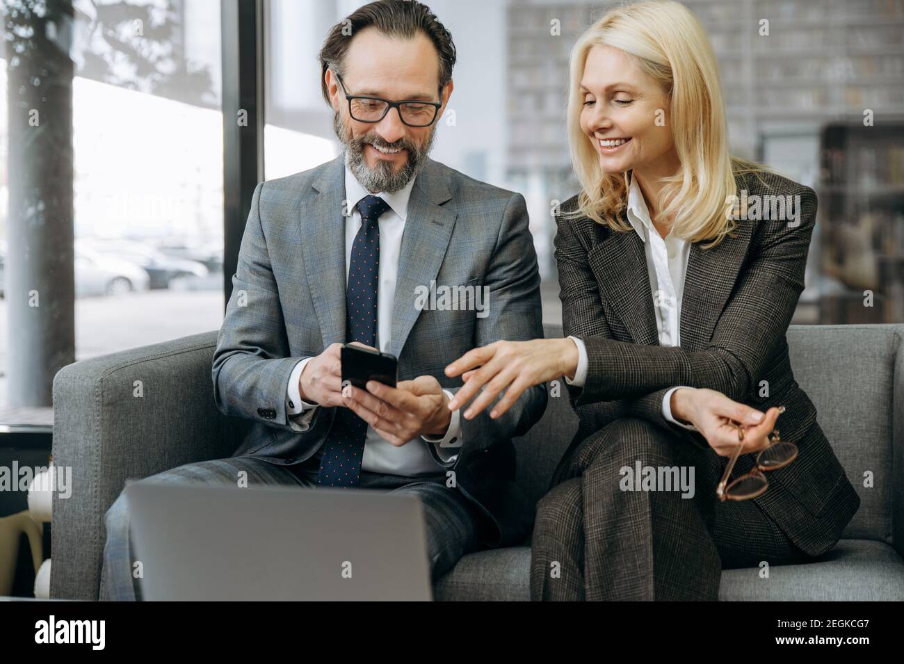Glückliche freundliche Geschäftspartner mit Smartphone, Surfen im Internet. Erfolgreiche Mitarbeiter in formellen Anzügen sitzen auf dem Sofa, machen eine Pause von der Arbeit, diskutieren Informationen Stockfoto