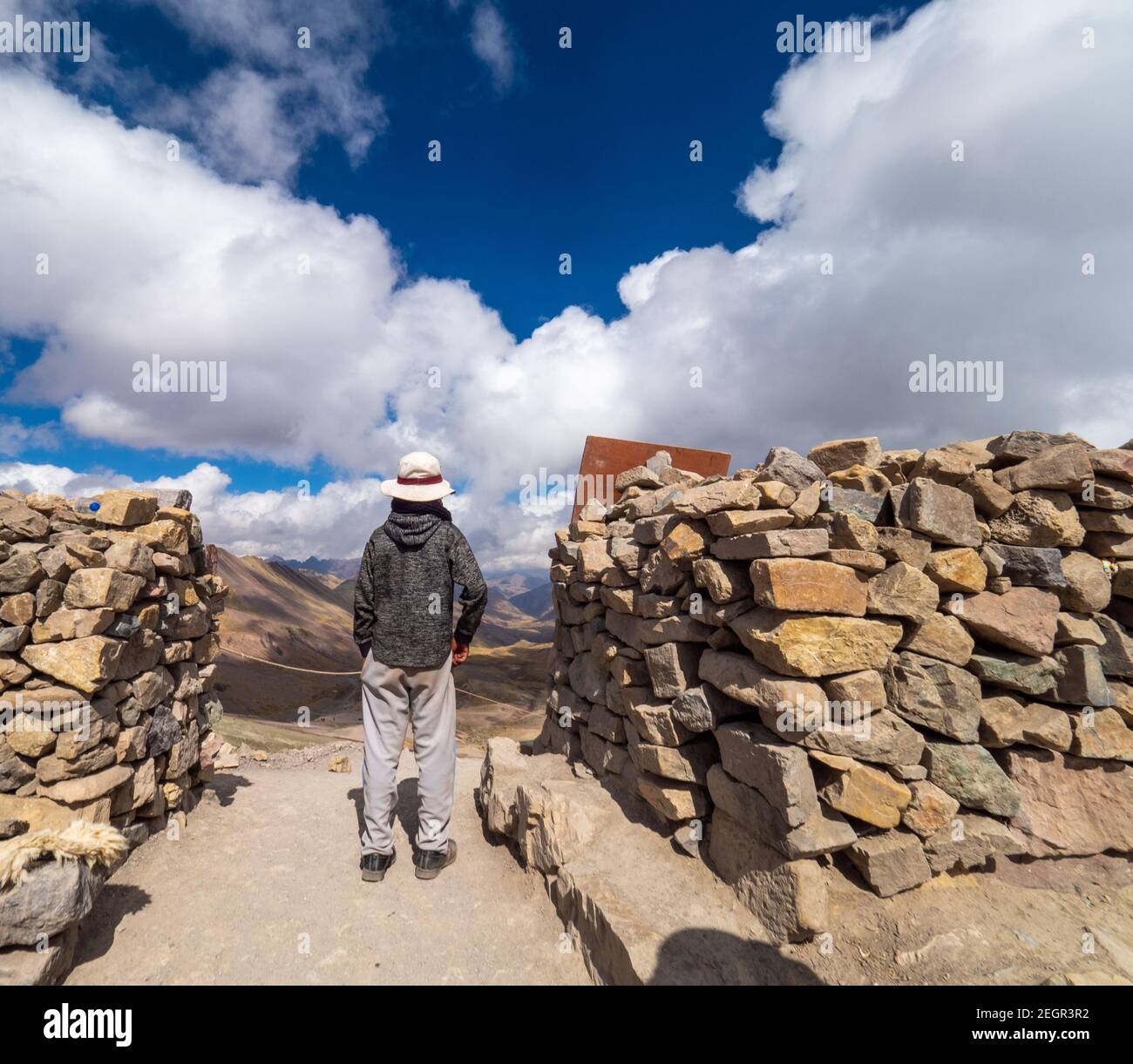 Peru, Vinicunca - 1. Oktober 2019 - junger Mann genießt den Blick von der Spitze des Berges neben Steinmauer, große weiße Wolken am Himmel Stockfoto