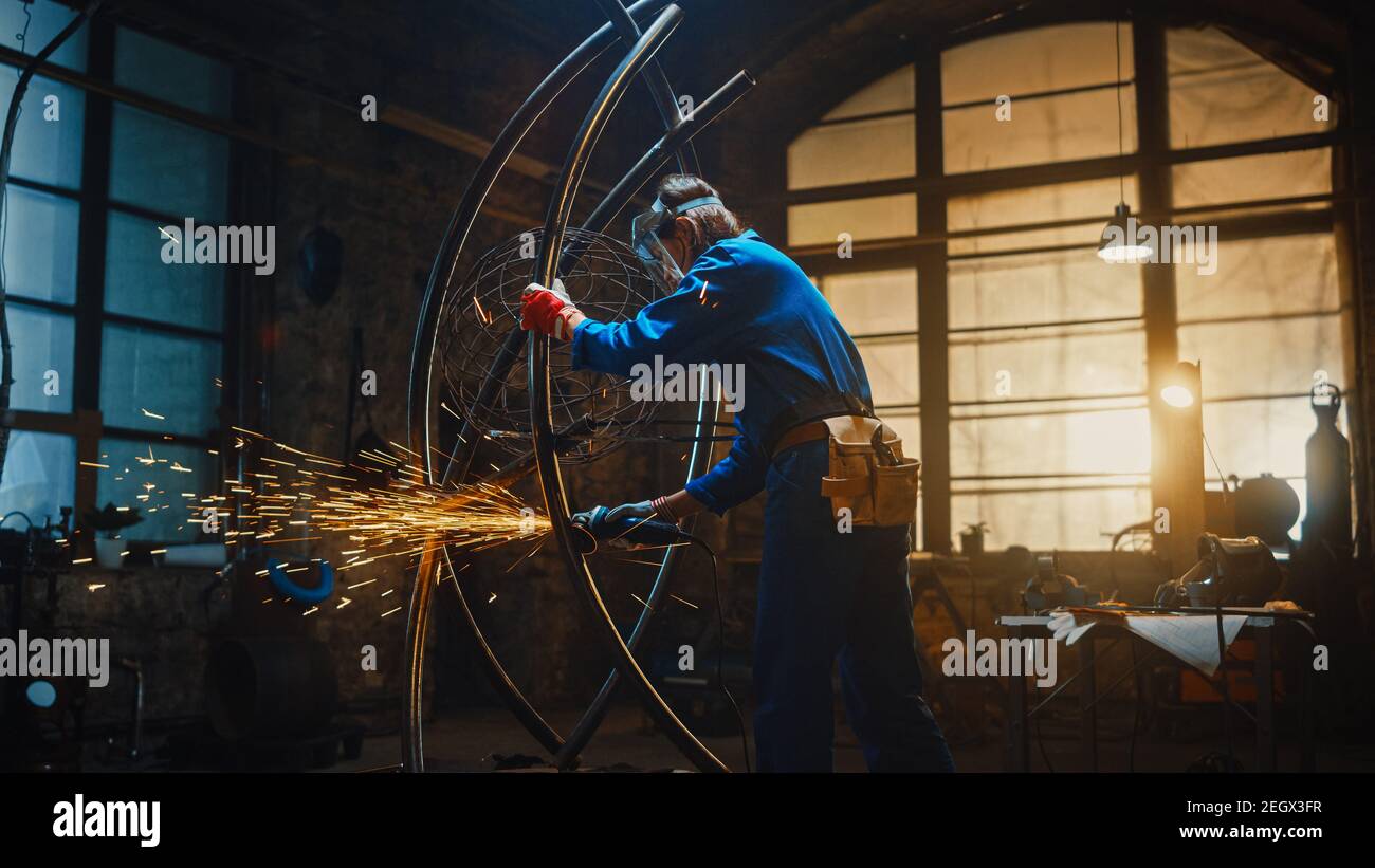 Junge zeitgenössische Künstlerin in Blue Jumpsuit ist Grinding eine Metallrohr Skulptur mit einem Winkel Grinder in einem Studio-Workshop. Befähigende Frau Stockfoto