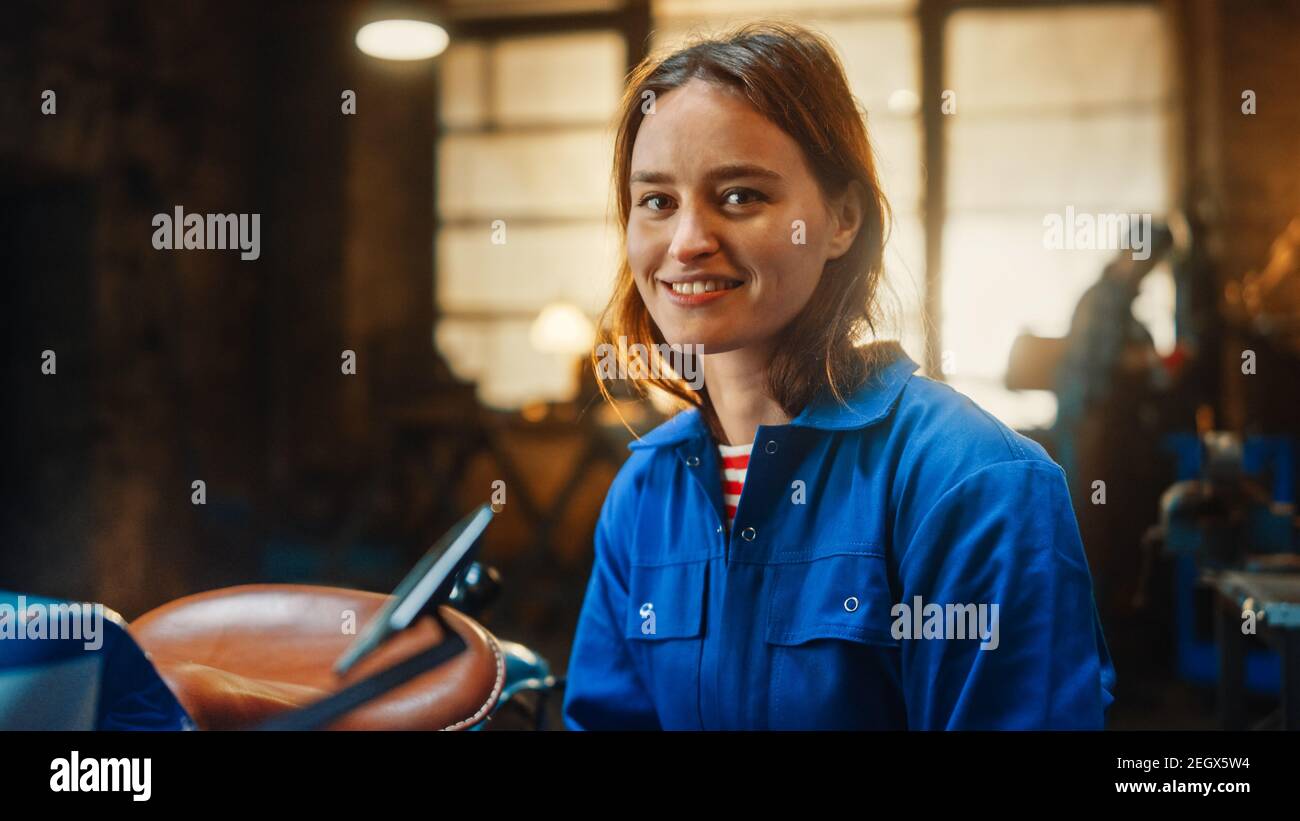 Junge schöne Ermächtigung Frau sanft Smiles an der Kamera. Authentischer Fabrikator trägt Arbeitskleidung in einer Metallwerkstatt. Stockfoto
