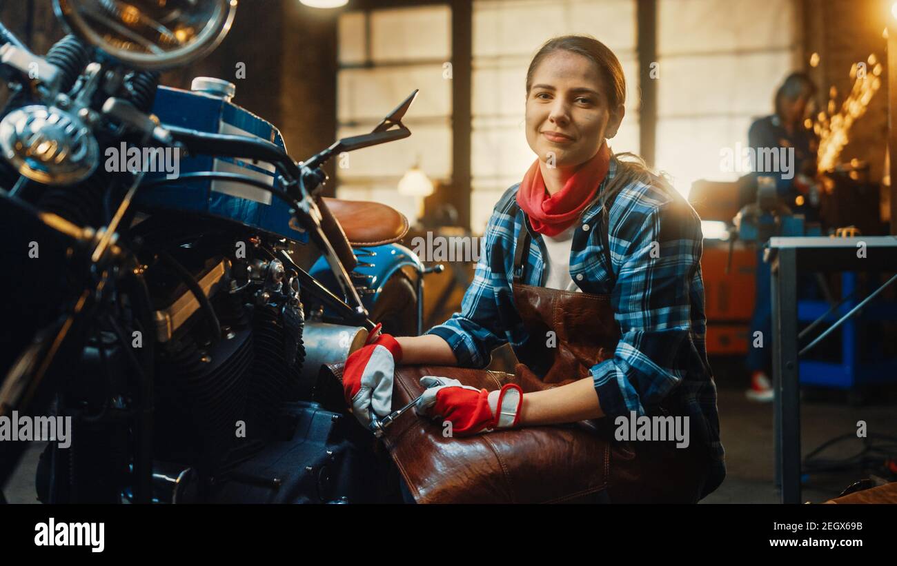 Junge schöne weibliche Mechaniker arbeitet an einem Custom Bobber Motorrad. Talentiertes Mädchen in einem karierten Hemd und einer Schürze. Sie smiles bei der Stockfoto