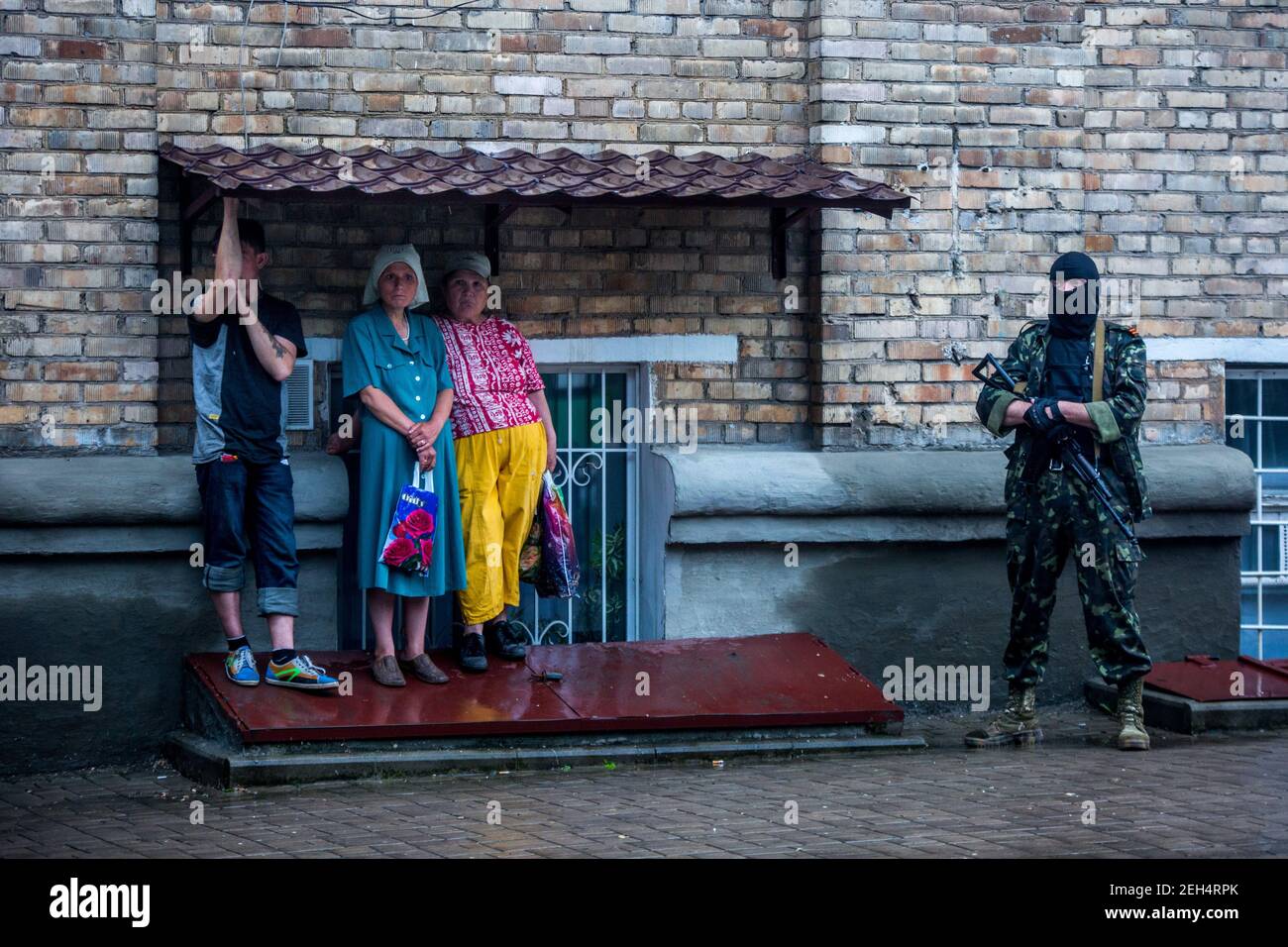 Michael Bunel / Le Pictorium - Ukraine Donbass-Krieg - 29/05/2014 - Ukraine / Donbass / Donezk - zwei Frauen und ein Mann, der vor dem Regen schützt, während ein Mann mit Gewehr und Kapuze neben ihnen steht. Nach den Kämpfen am Flughafen Donezk sollen die Pro-Russen einen Supermarkt ausgeraubt haben. Die Männer des Bataillons Wostok kamen ein paar Tage früher an und wären an diesem Tag gekommen, um die Verantwortlichen zu verhaften. Zu dieser Zeit wurde eine Operation durchgeführt, um sie aus der OGA, dem Gebäude der regionalen Staatsverwaltung, zu entfernen, die seit Beginn des separatistischen Aufstands besetzt war. Da die b Stockfoto