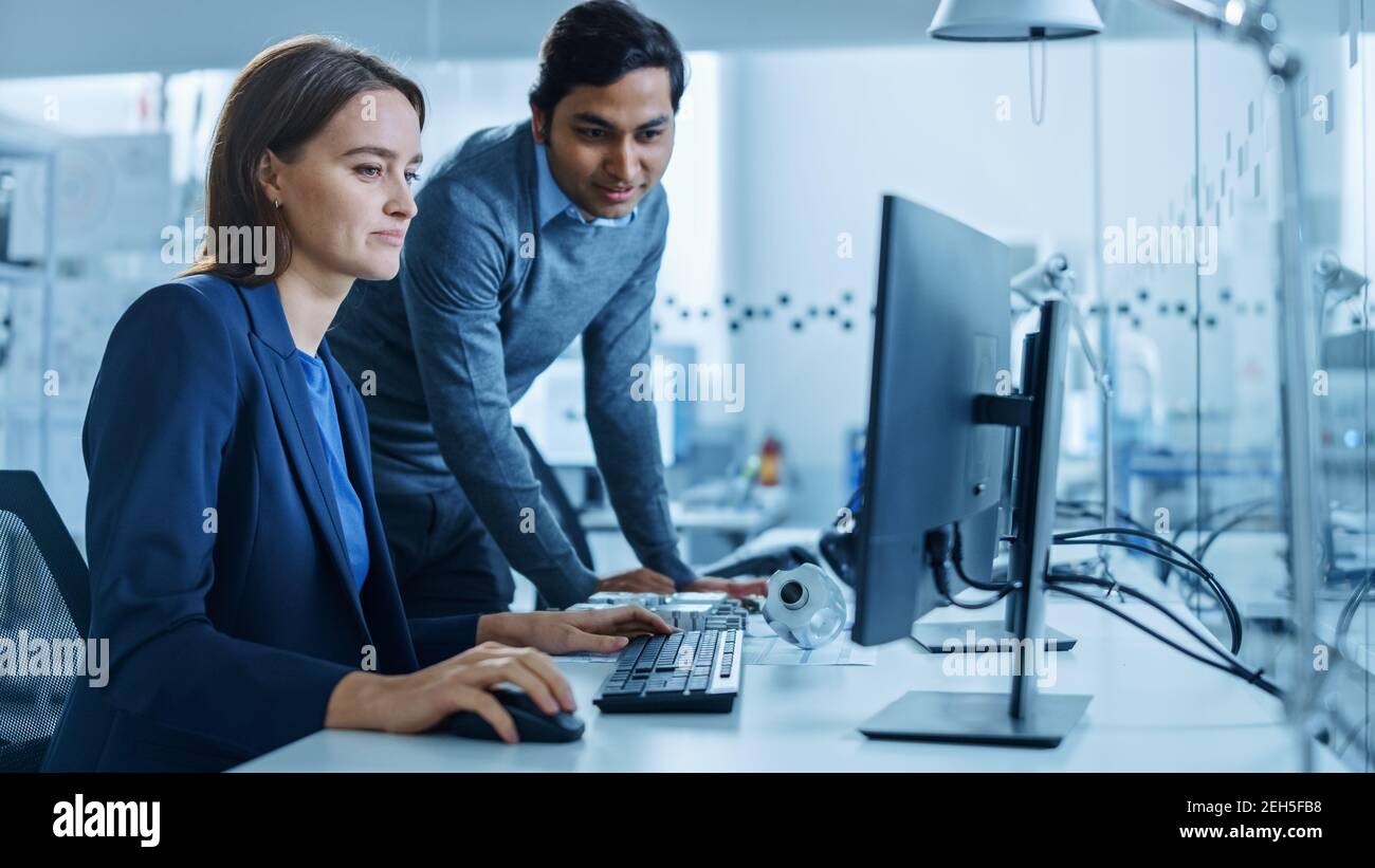 Moderne Fabrik Büro: Männliche Projektleiterin spricht mit einer weiblichen Industrieingenieurin, die auf Computer arbeitet. Professionelle Teamarbeit, Junge Spezialisten Stockfoto