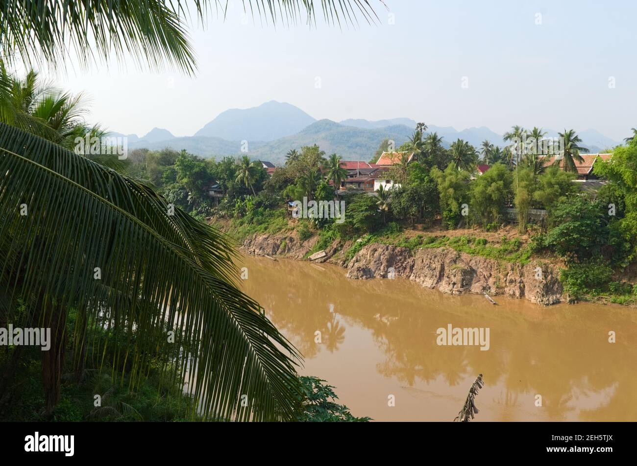 Blick auf den Mekong durch Palmenblätter. Orangefarbenes Dach des buddhistischen Tempels und Berge im Hintergrund. Traditionelle Landschaft in Laos. Stockfoto