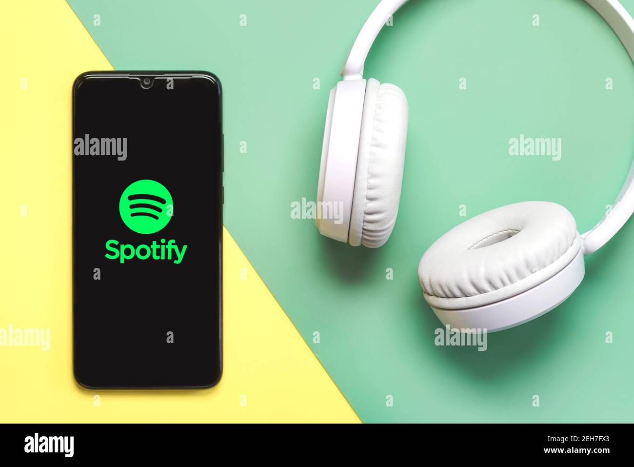 Spotify-Logo auf schwarzem Smartphone-Bildschirm mit weißen Kopfhörern Auf einem grünen und gelben Hintergrund Stockfoto