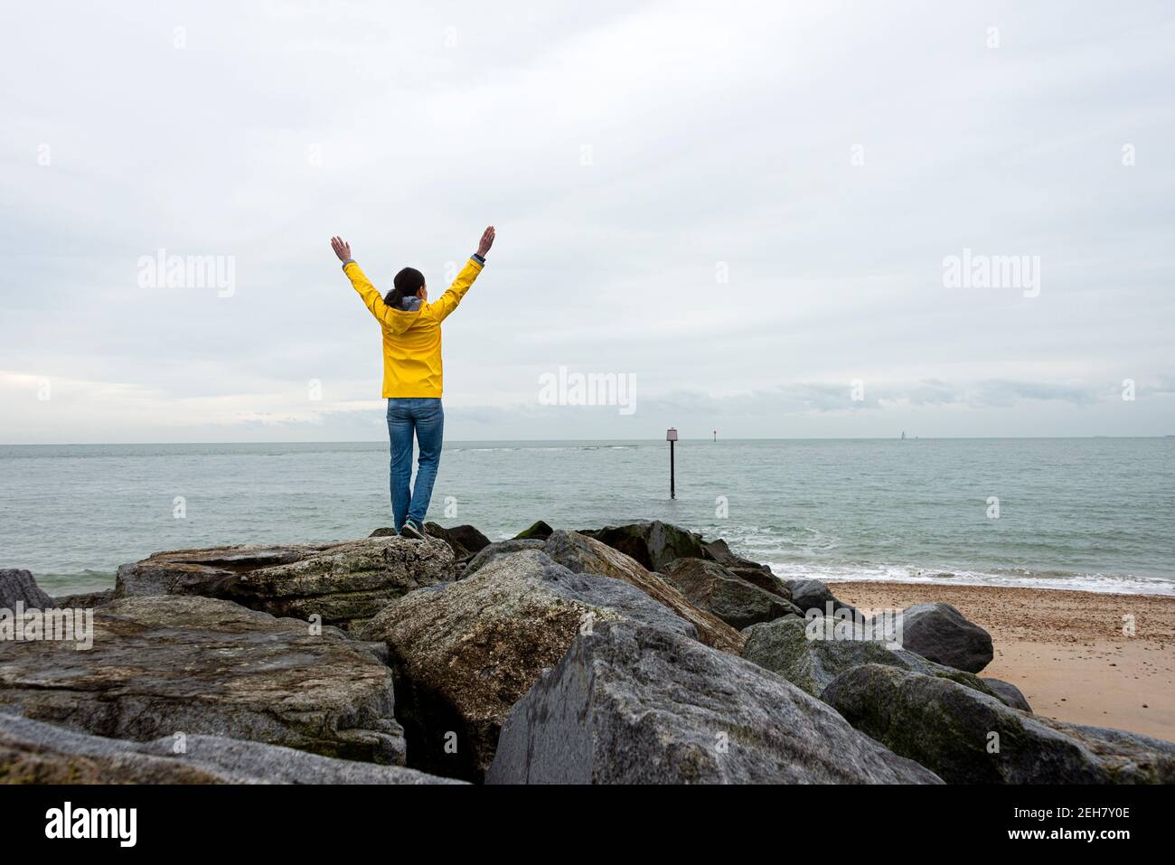 Rückansicht einer Frau, die an einem grau bewölkten Tag auf Felsen mit erhobenen Armen steht und auf das Meer blickt. Stockfoto