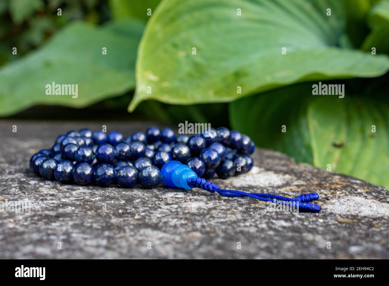 Blaue Gebetsperlen (Lapislazuli) auf Stein mit grünen Blättern im Hintergrund, Nahaufnahme. Achtsamkeit und Meditationszubehör. Stockfoto