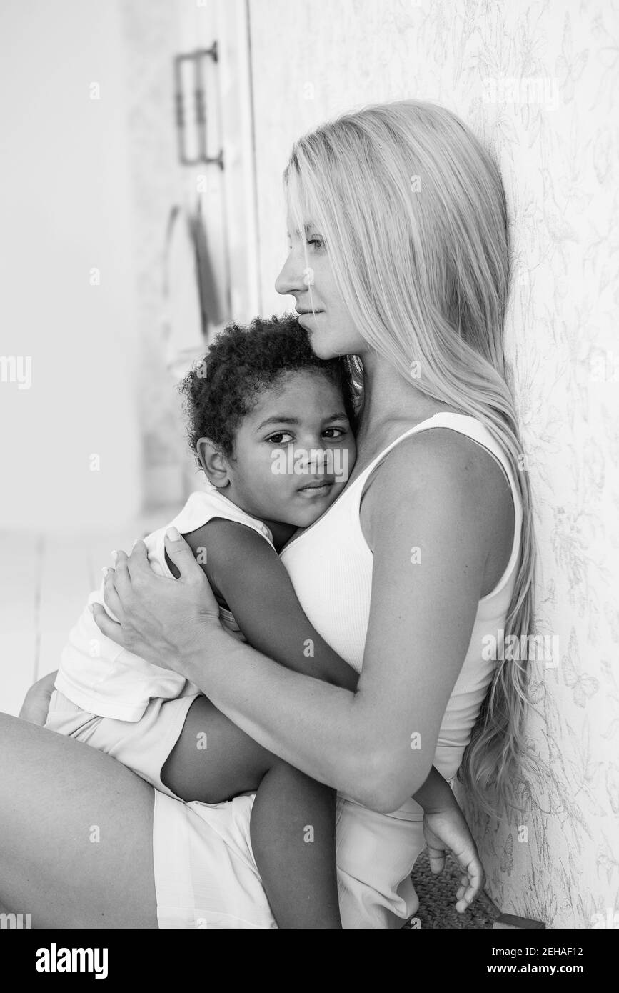 Porträt einer jungen blonden kaukasischen Mutter hält, umarmt kleinen afroamerikanischen Sohn in den Armen. Der Junge drückte gegen die Brust der Frau. Zärtlichkeit Stockfoto