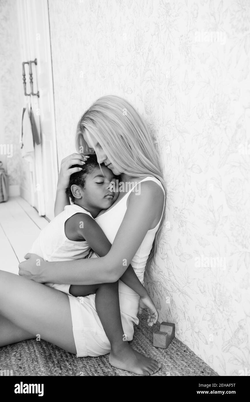 Porträt einer jungen blonden kaukasischen Mutter hält, umarmt kleinen afroamerikanischen Sohn in den Armen. Der Junge drückte gegen die Brust der Frau. Zärtlichkeit Stockfoto