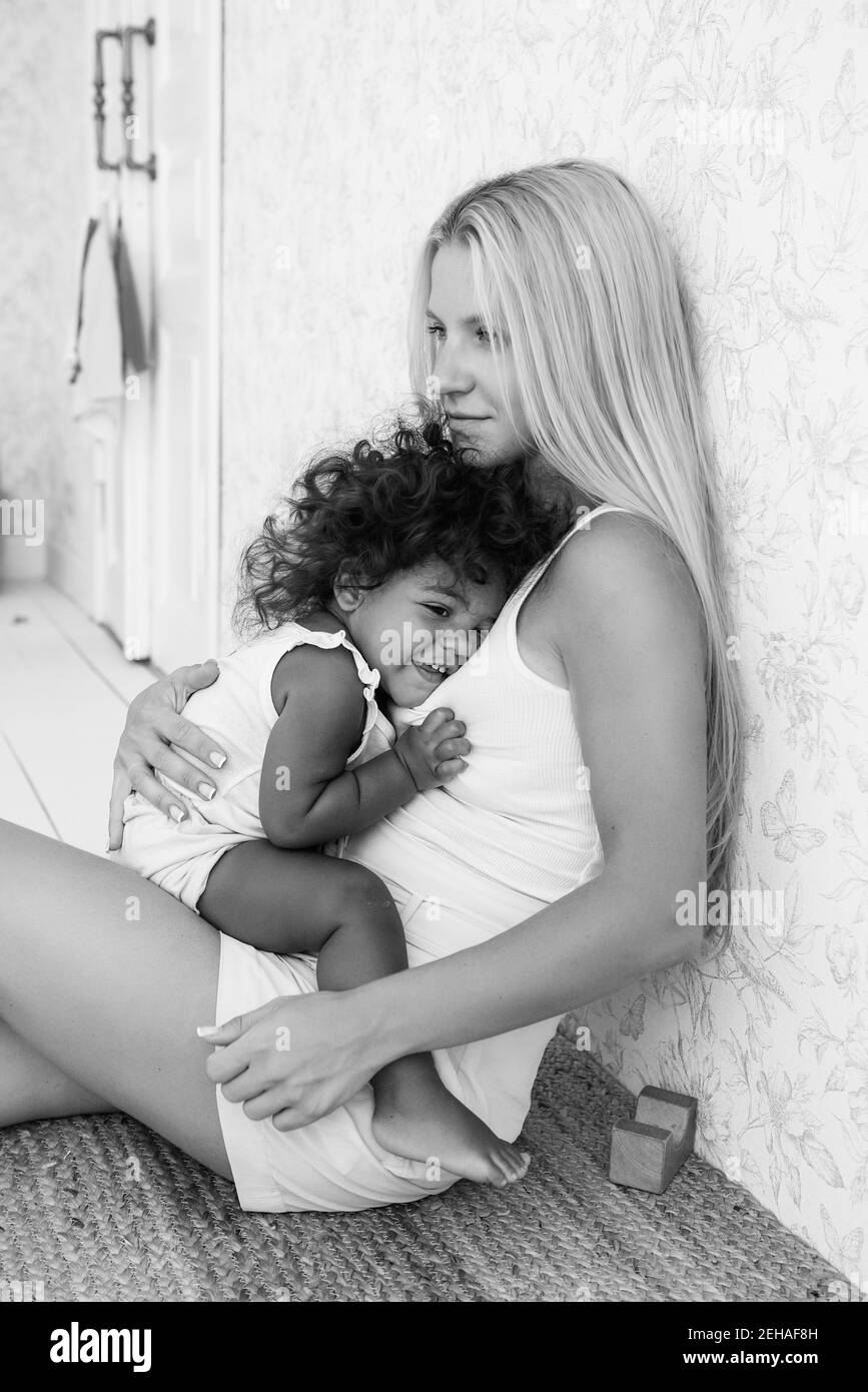 Porträt der jungen blonden kaukasischen Mutter hält Umarmung kleine afroamerikanische Tochter in den Armen. Mädchen drückte gegen die Brust der Frau. Zärtlichkeit Stockfoto