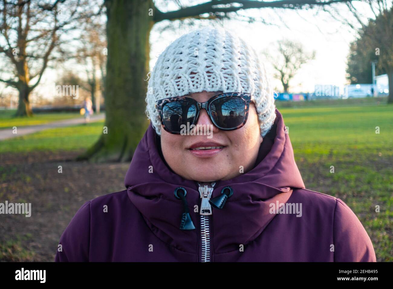 Porträt einer asiatischen Dame in einem Park mit warmem Wintermantel, Hut und Sonnenbrille im späten Nachmittagssonne an einem Tag im Februar 2021. Stockfoto